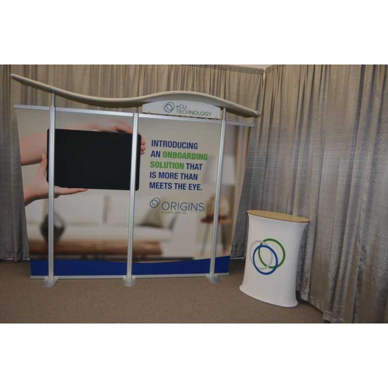 The 10ft Timberline Wave Canopy Tapered Kit 5 by Affordable Exhibit Displays features an SEG fabric banner—INTRODUCING AN ONBOARDING SOLUTION THAT IS MORE THAN MEETS THE EYE—a small counter with overlapping circles, and beige curtains.