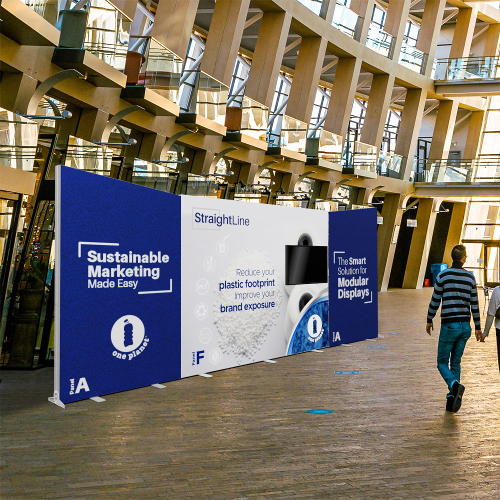 A person passes a 20ft StraightLine AFA Kit by Affordable Exhibit Displays showing “Sustainable Marketing Made Easy” and “Reduce your plastic footprint to improve your brand exposure” in a bright, modern building with tall windows.