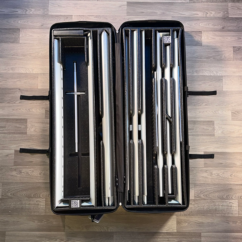 A large black carrying case from Affordable Exhibit Displays holds components for the 20ft BrightLine MKKM Kit, including neatly organized metal and carbon fiber rods separated by foam dividers on a wooden floor.
