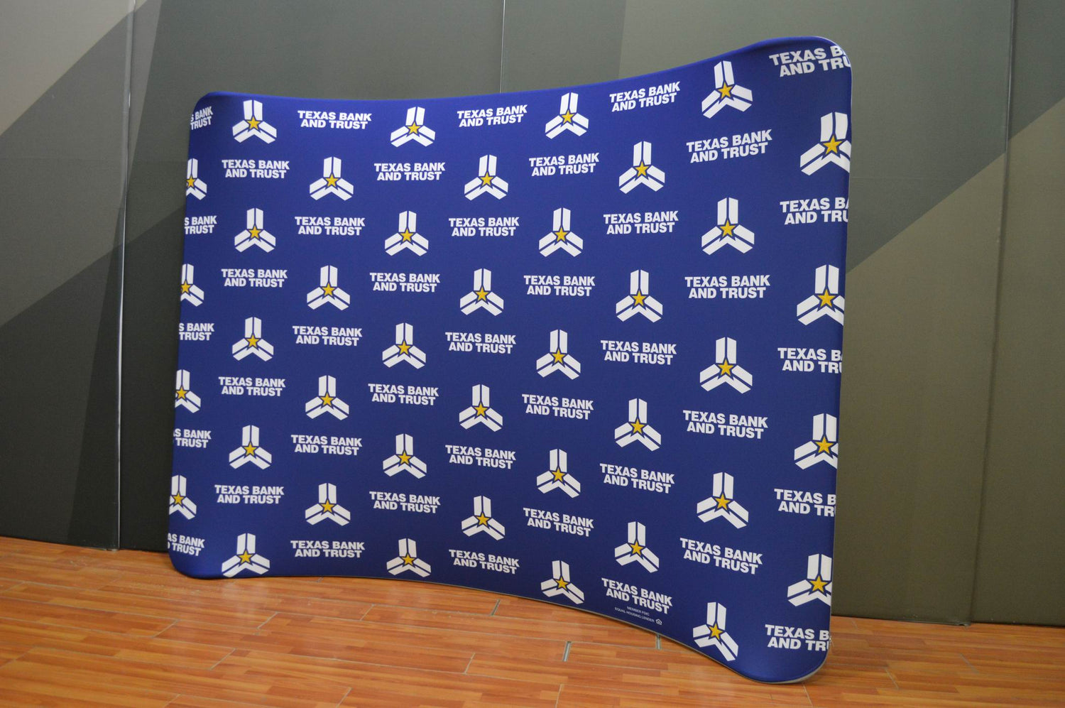 A blue backdrop with repeated Texas Bank and Trust logos and text stands on a wooden floor against a gray and green wall.